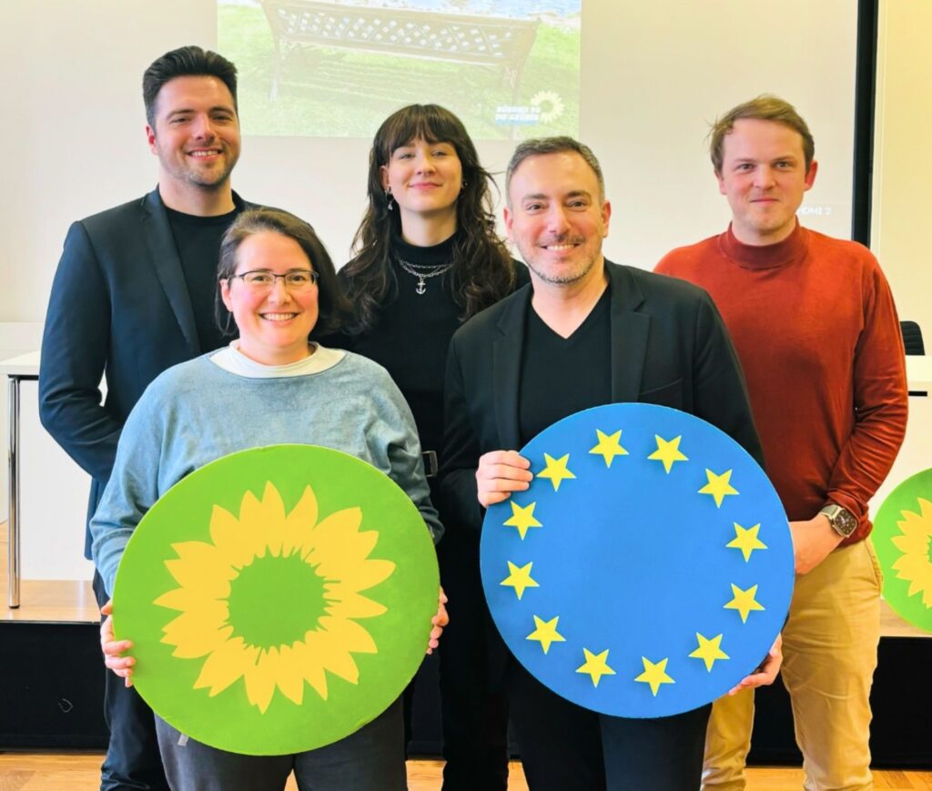 Gruppenbild der Kandidierenden für das Europaparlament, 5 Menschen lächeln,
2 halten runde Schilder hoch - eine gelbe Sonnenblume auf grünem Hintergrund und ein blauer Kreis mit gelben Sternen.
Ein Logo der Grünen und eine Europafahne.
